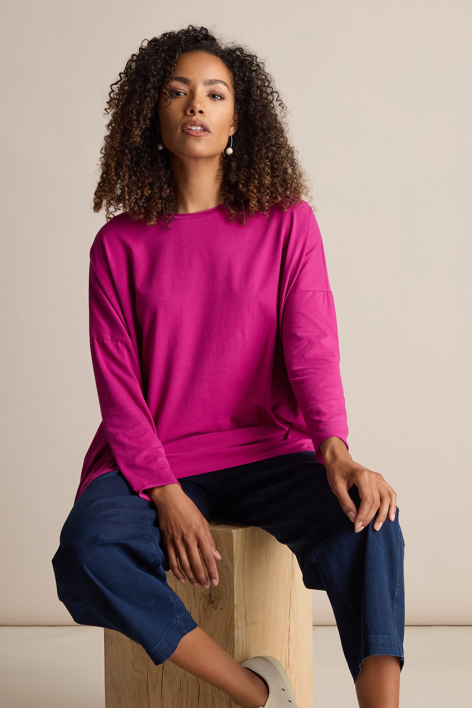 Person with curly hair wearing a vibrant pink Cotton Jersey Relaxed Top and blue pants, sitting on a wooden block against a plain background.