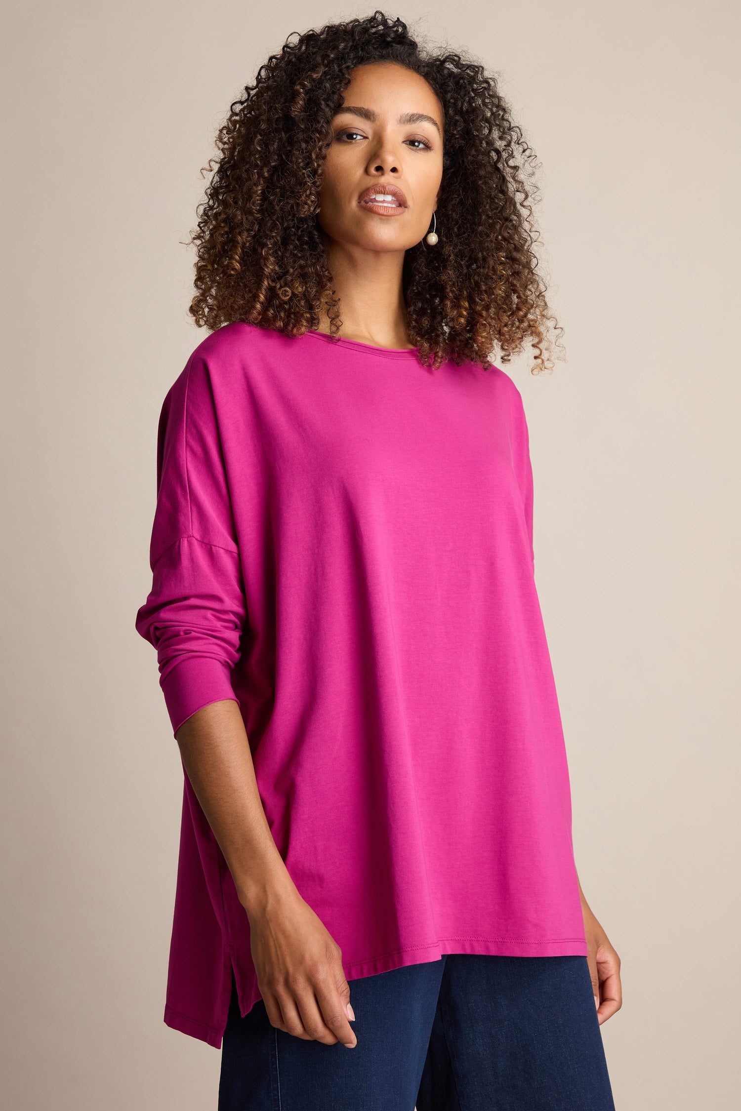 A person with curly hair stands against a neutral background wearing a vibrant pink Cotton Jersey Relaxed Top and dark pants, perfect for the Autumn season.