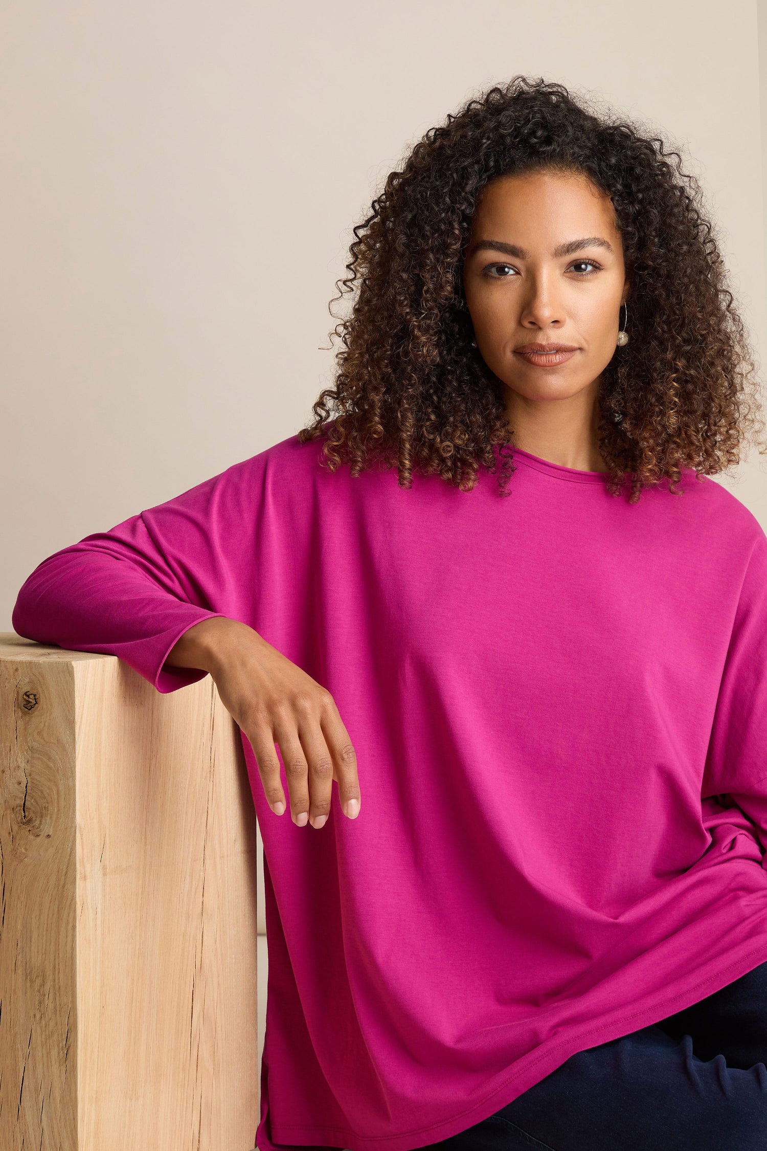 A person with curly hair wearing a bright pink Cotton Jersey Relaxed Top with long sleeves rests one arm on a wooden surface while looking at the camera.