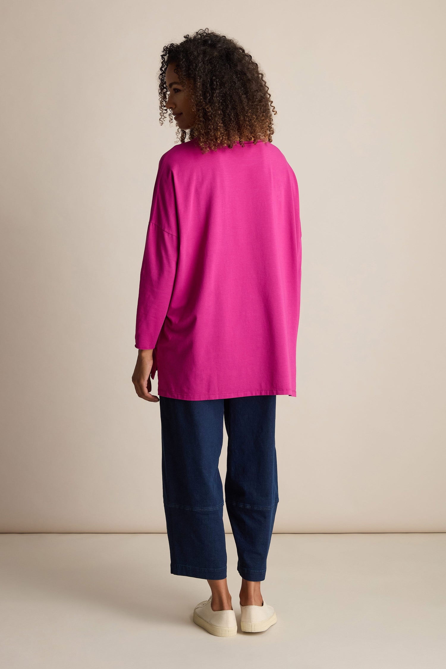 A person with curly hair stands facing away, wearing a bright pink Cotton Jersey Relaxed Top, dark blue pants, and beige shoes against a plain beige background.