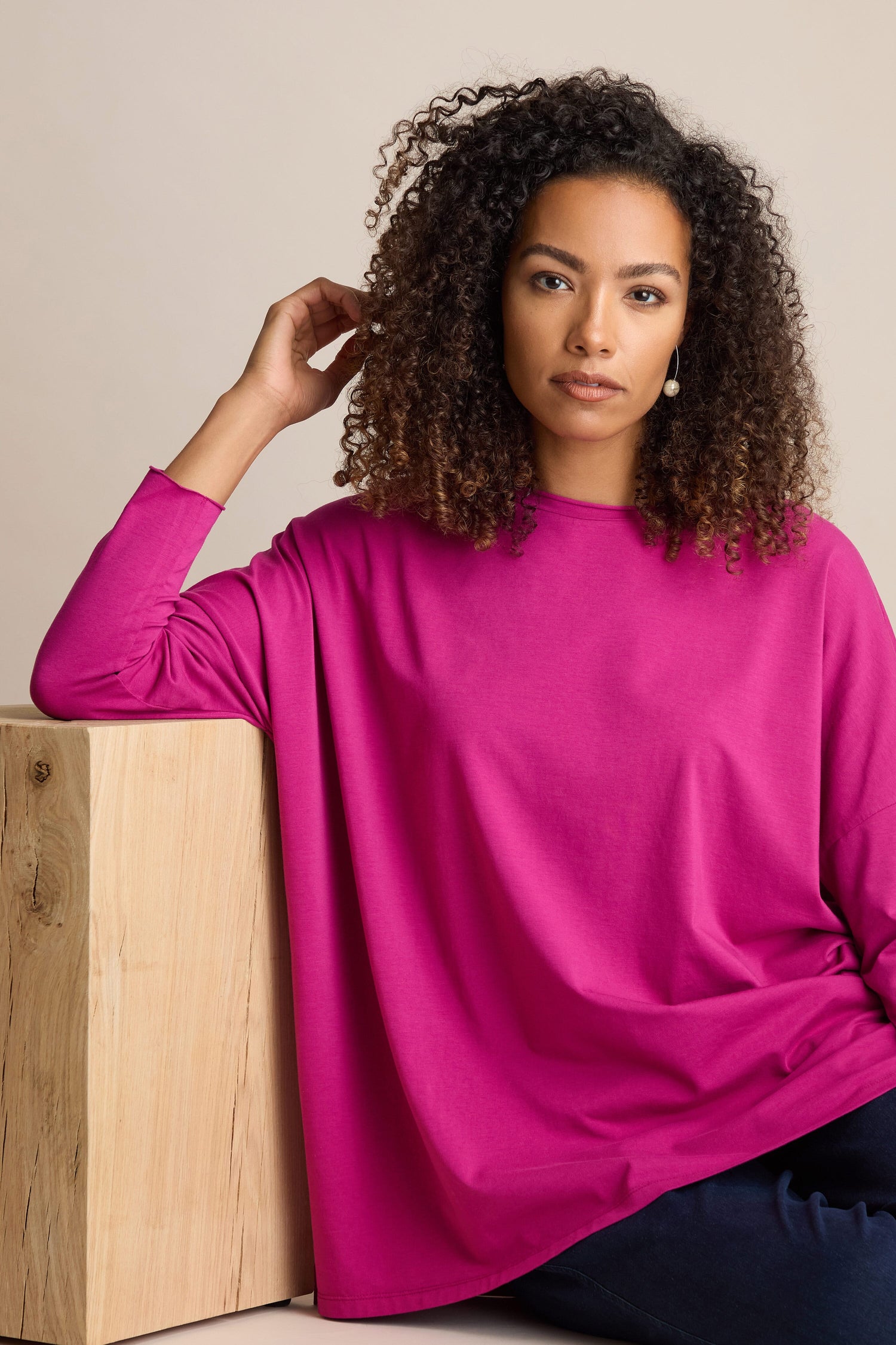 A woman with curly hair, wearing a Cotton Jersey Relaxed Top in bright pink, leans against a light-colored wooden block, posing confidently for the camera.