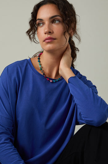 A woman with curly hair wears a blue Cotton Jersey Relaxed Top and a colorful beaded necklace, sitting with her hand on her neck and looking to the side.