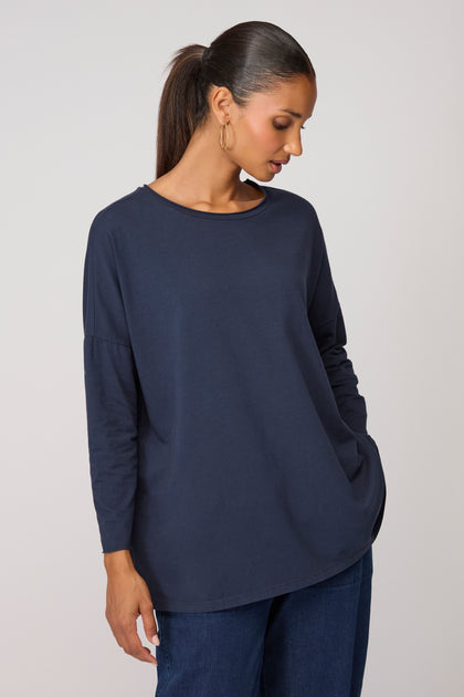 Model wears the Cotton Jersey Relaxed Top in navy blue with blue jeans, standing casually with one hand in her pocket and gazing down—an effortless wardrobe staple set against a plain background.