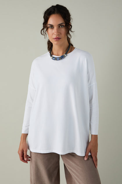 A woman stands against a plain light background wearing the Cotton Jersey Relaxed Top, styled for comfort, paired with beige pants and a chunky necklace.