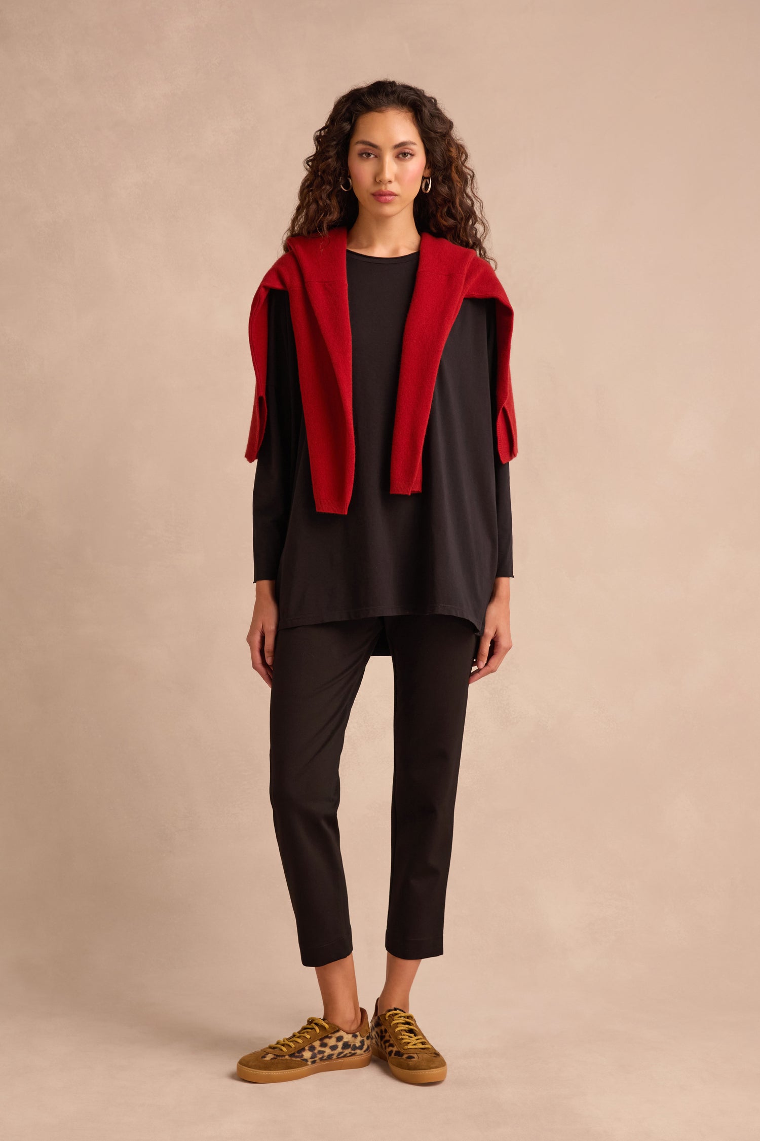 Woman standing against a beige background, wearing the Black Cotton Jersey Relaxed Top and matching relaxed-fit pants, a red sweater draped over her shoulders, and brown leopard-print sneakers.