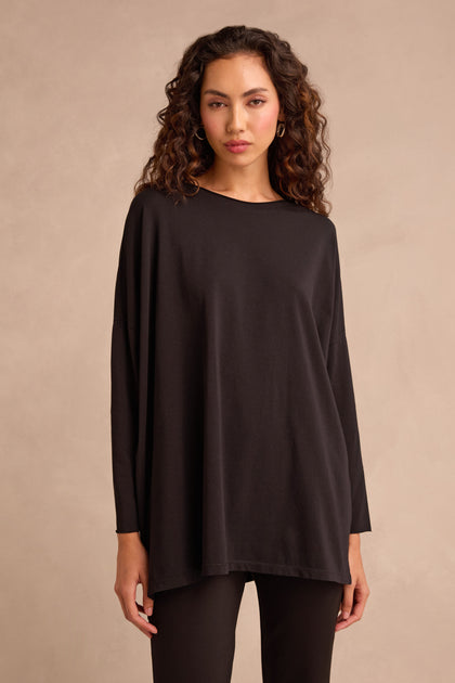 A woman with wavy brown hair wears the Black Cotton Jersey Relaxed Top paired with black pants featuring a boxy silhouette, standing against a neutral beige background.