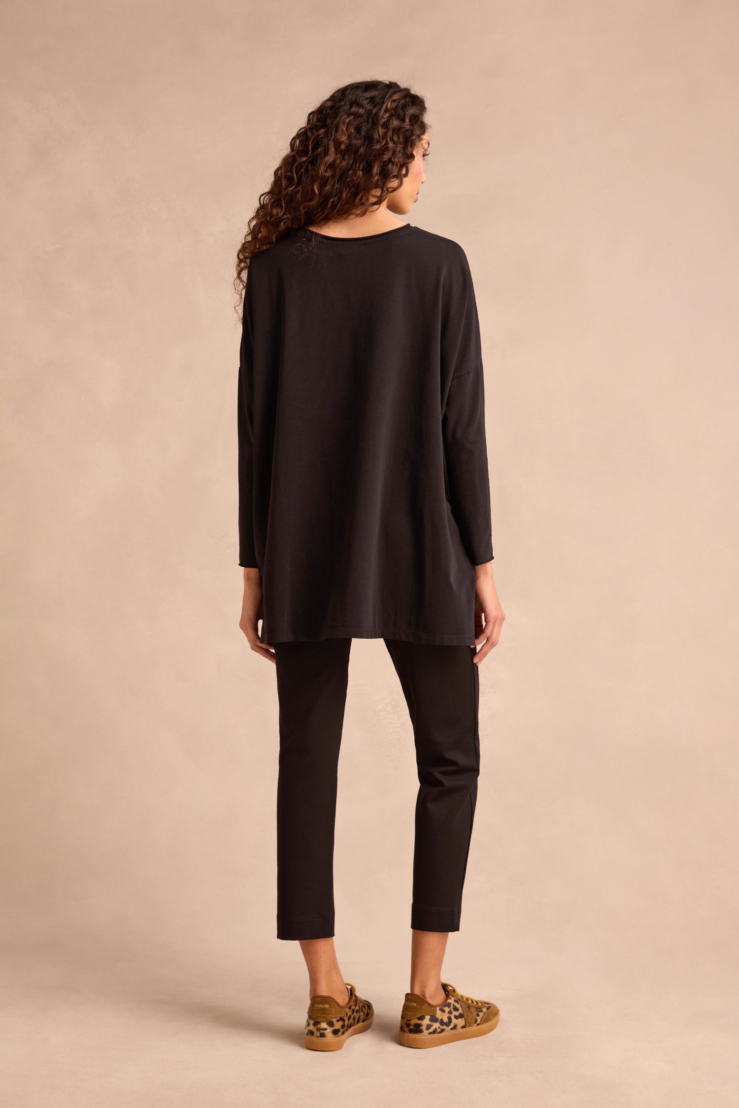 A person with long curly hair is standing, facing away, wearing the Black Cotton Jersey Relaxed Top, black pants, and leopard print shoes against a neutral beige background.