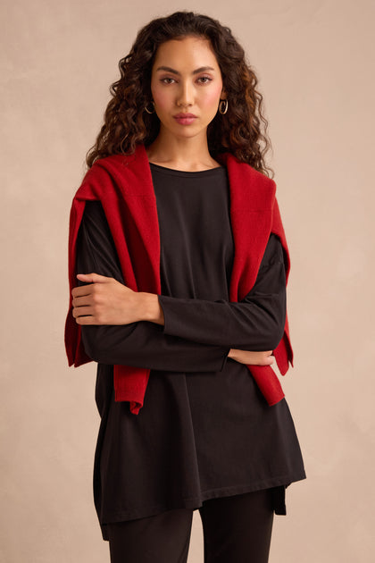 A woman with long curly hair stands against a beige background with her arms crossed, wearing the Black Cotton Jersey Relaxed Top and matching pants, with a red sweater draped over her shoulders.