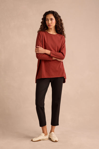 A woman stands against a beige background, wearing the Conker Cotton Jersey Relaxed Top—a rust-colored long-sleeve cotton jersey in a relaxed silhouette—paired with black ankle-length pants and white sneakers.