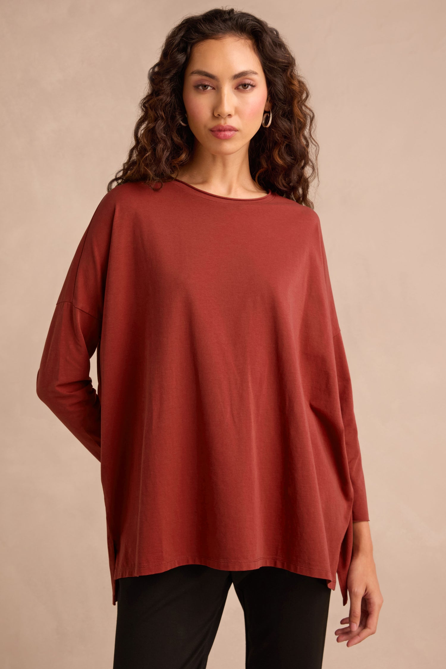 Woman with long curly hair wearing the Conker Cotton Jersey Relaxed Top—a women’s oversized, loose-fitting rust-colored top crafted from soft cotton jersey—and black pants, standing against a plain light background. The relaxed silhouette ensures comfort and effortless style.
