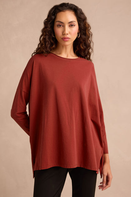 Woman with long curly hair wearing the Conker Cotton Jersey Relaxed Top—a women’s oversized, loose-fitting rust-colored top crafted from soft cotton jersey—and black pants, standing against a plain light background. The relaxed silhouette ensures comfort and effortless style.
