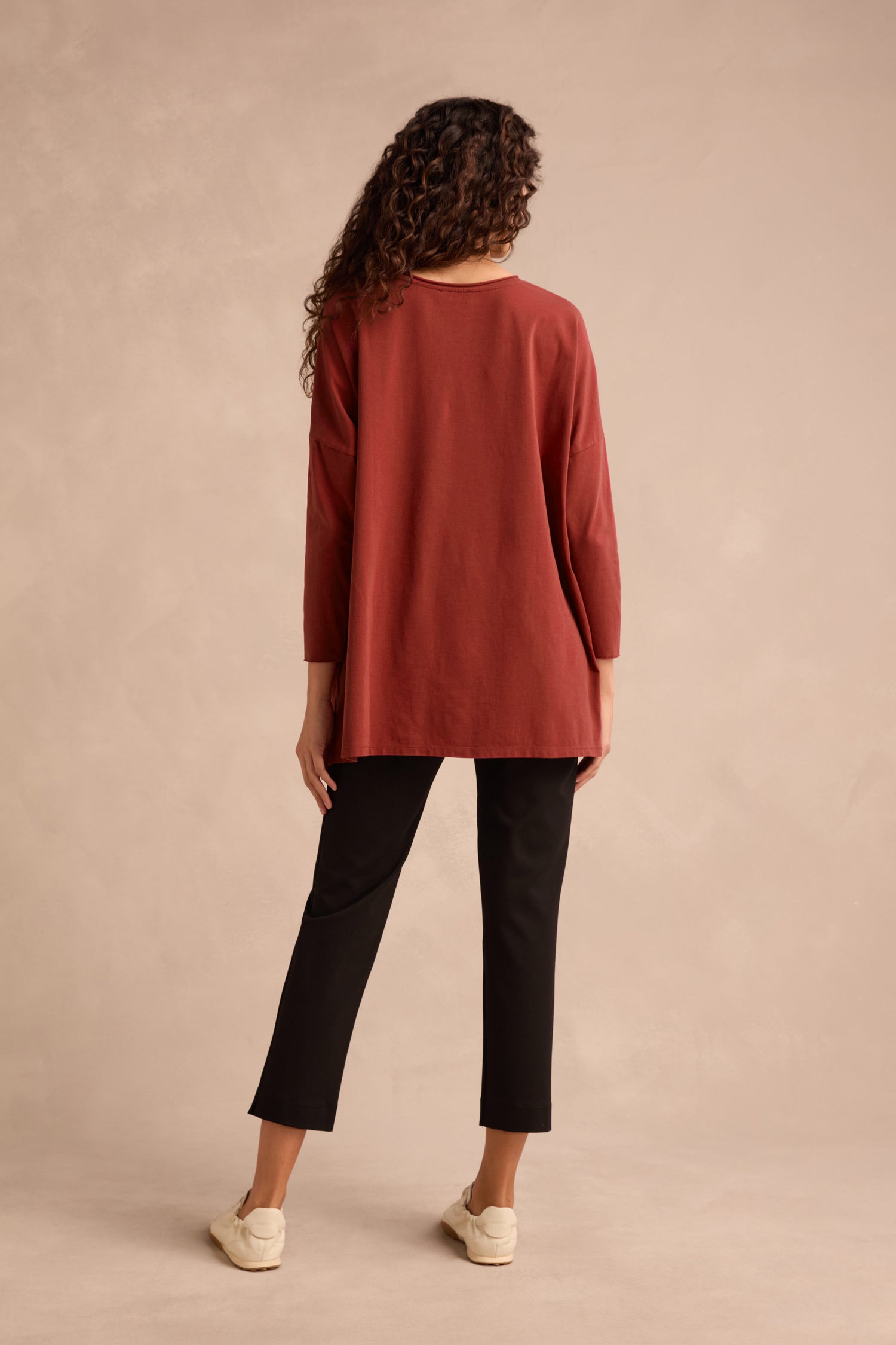 A person stands facing away, wearing the Conker Cotton Jersey Relaxed Top in a relaxed silhouette with black pants and cream shoes, against a beige background.