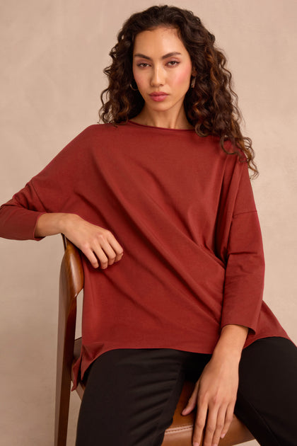 A woman with long, curly hair sits on a wooden chair, wearing the Conker Cotton Jersey Relaxed Top in rust and black pants against a neutral background.