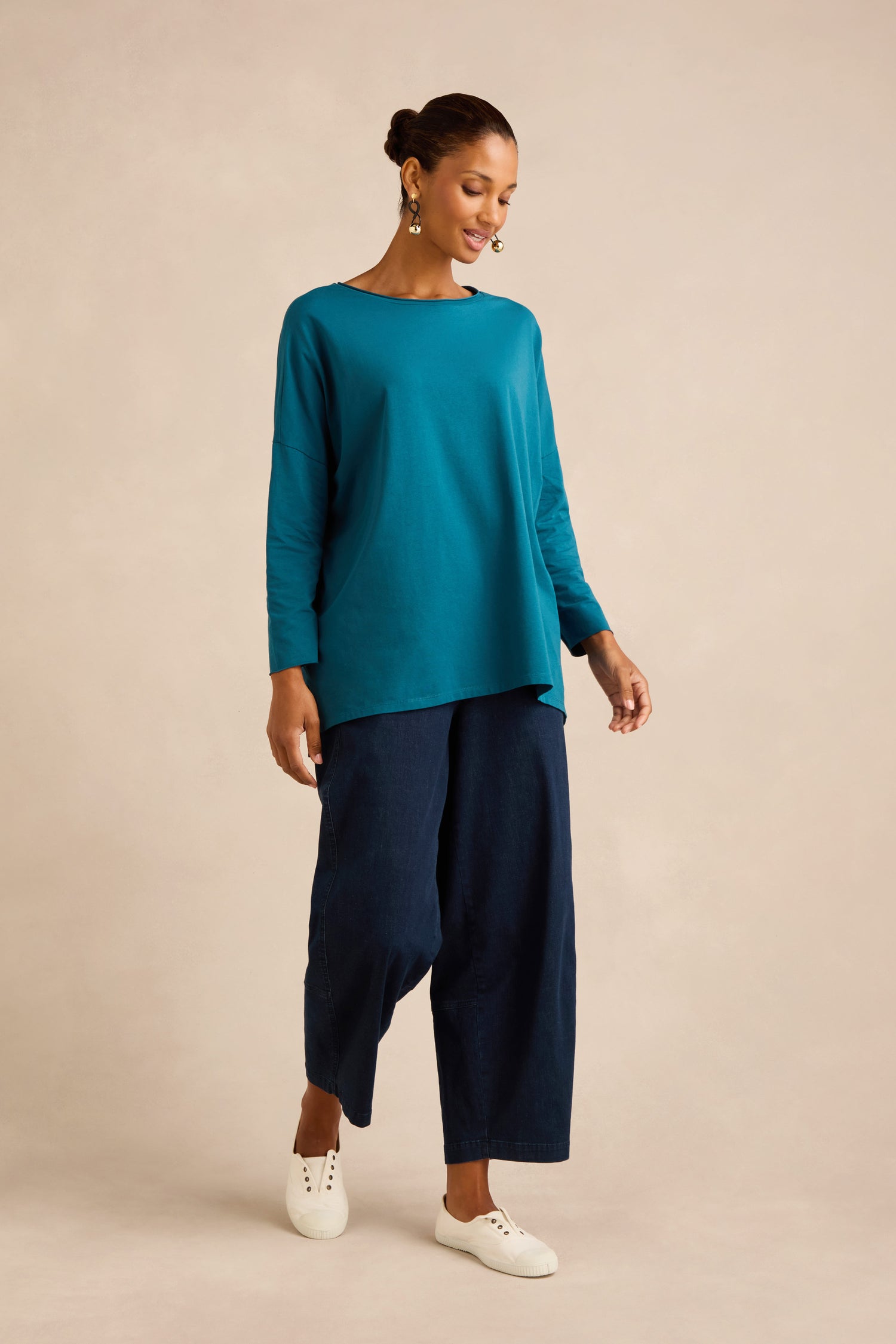 A woman stands against a neutral background wearing the Cotton Jersey Relaxed Top in a boxy silhouette, paired with wide-leg dark blue pants and white sneakers.