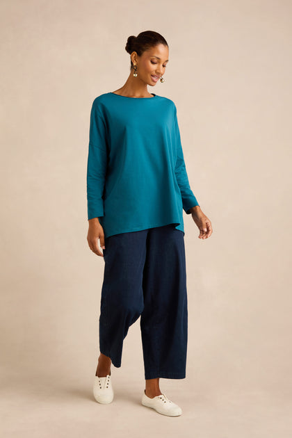 A woman stands against a neutral background wearing the Cotton Jersey Relaxed Top in a boxy silhouette, paired with wide-leg dark blue pants and white sneakers.