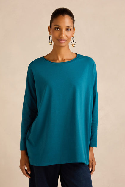 A woman wearing the Cotton Jersey Relaxed Top in teal, featuring a loose fit and boxy silhouette, paired with dark pants. She stands against a plain beige background, looking at the camera.