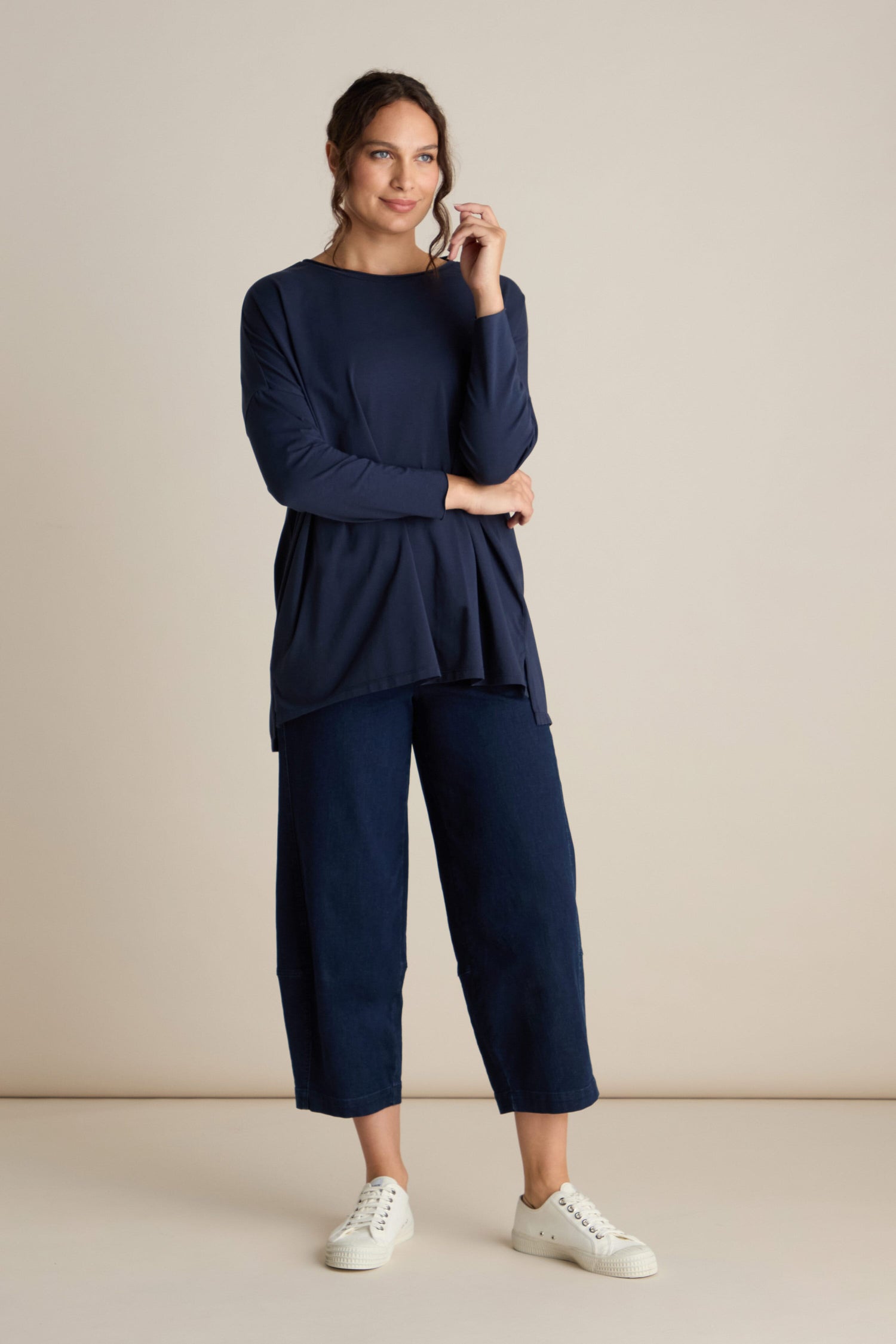 A woman is wearing a luxurious Cotton Jersey Relaxed Top in dark blue, paired with matching wide-leg pants and white sneakers. She stands against a plain beige background with her left hand raised near her chin, effortlessly showcasing this elevated basic piece.