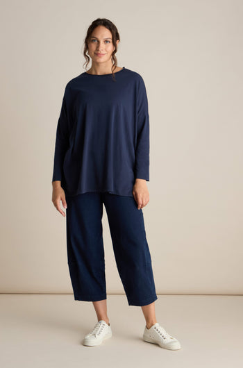 A person clad in a navy blue Cotton Jersey Relaxed Top, dark cropped pants, and white sneakers stands against a plain beige background. This elevated basic piece effortlessly merges comfort with style.