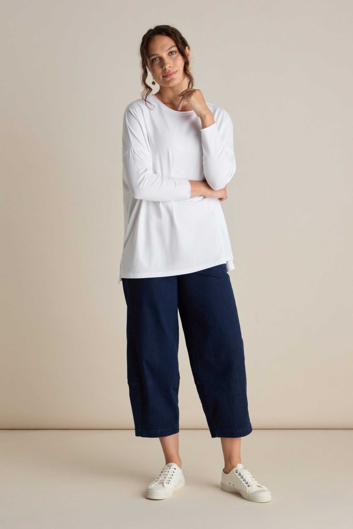 A person stands against a plain background, wearing an elevated basic piece: the Cotton Jersey Relaxed Top, paired with dark blue wide-leg pants and white sneakers. They look at the camera, one hand touching their face.