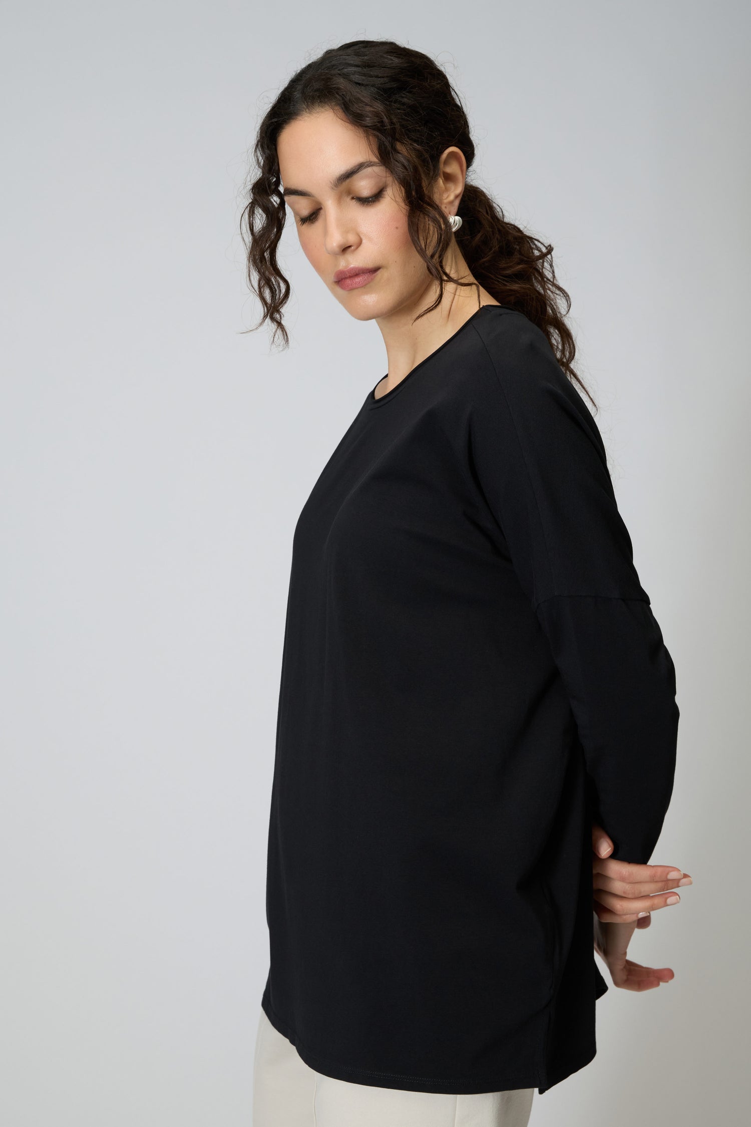 A woman with curly hair stands in side profile against a plain background, looking down with hands behind her back, wearing the Cotton Jersey Relaxed Top and light pants.