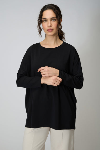 A woman with curly hair wears the Cotton Jersey Relaxed Top and light-colored pants against a plain, light background—the ideal wardrobe essential for effortless style.