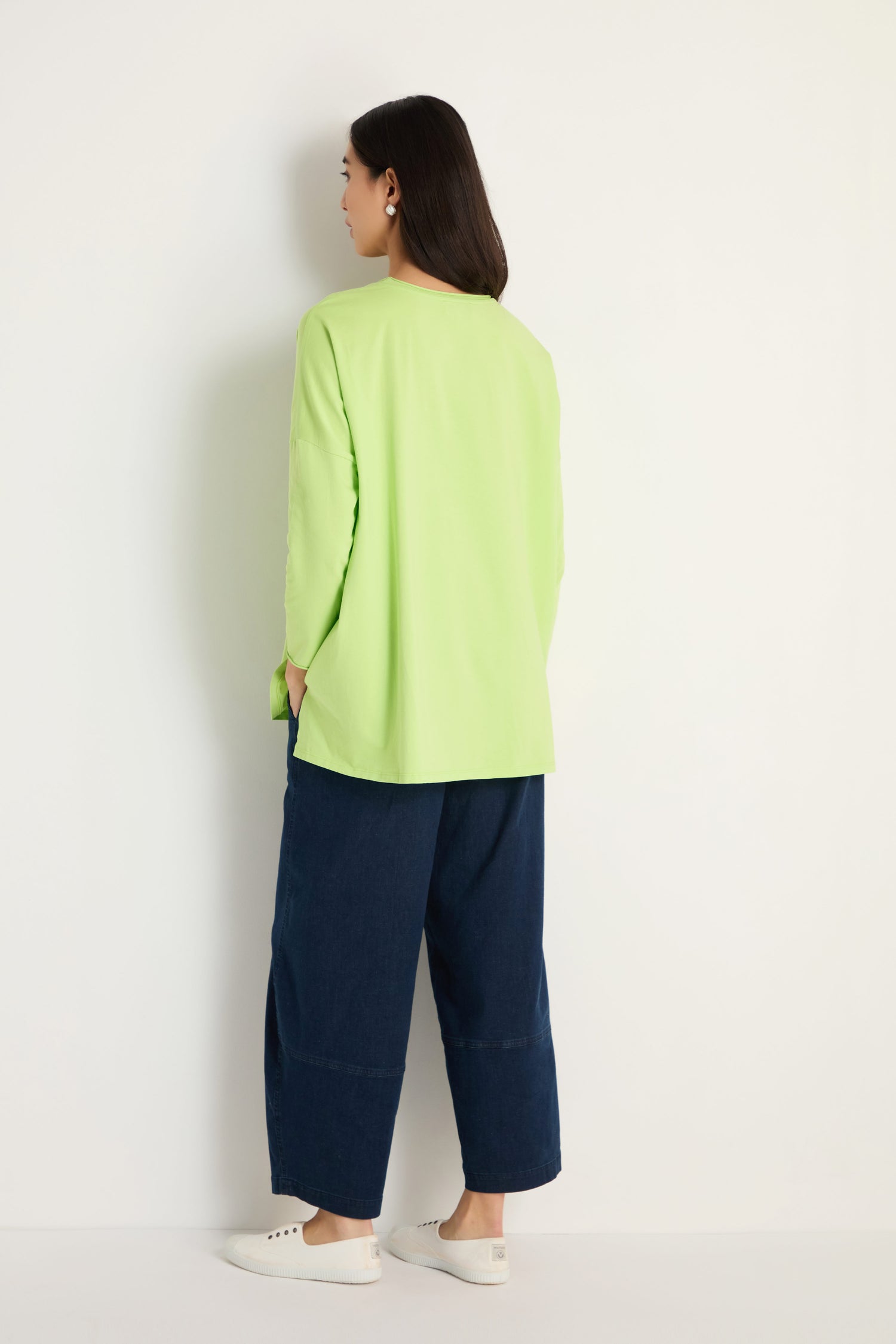 A person stands facing a white wall, wearing the Cotton Jersey Relaxed Top in light green, wide dark blue pants, and white shoes.