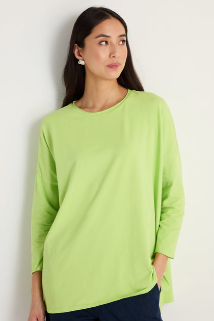 A woman in a relaxed silhouette, wearing the Cotton Jersey Relaxed Top in lime green with dark pants, stands against a plain light wall and looks to the side.