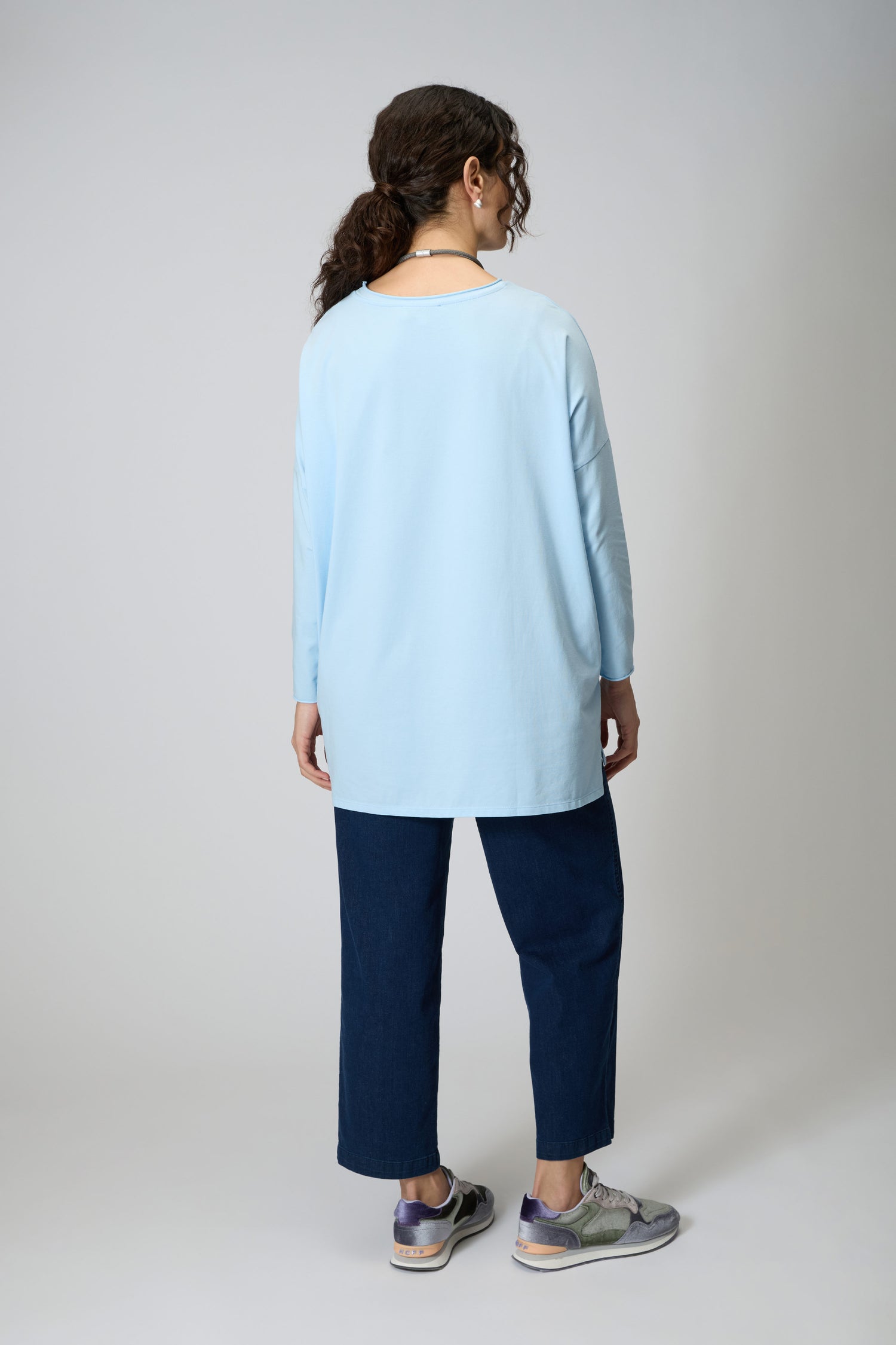 A person with curly hair, wearing a light blue Cotton Jersey Relaxed Top, dark blue pants, and sneakers stands facing away from the camera against a plain background.