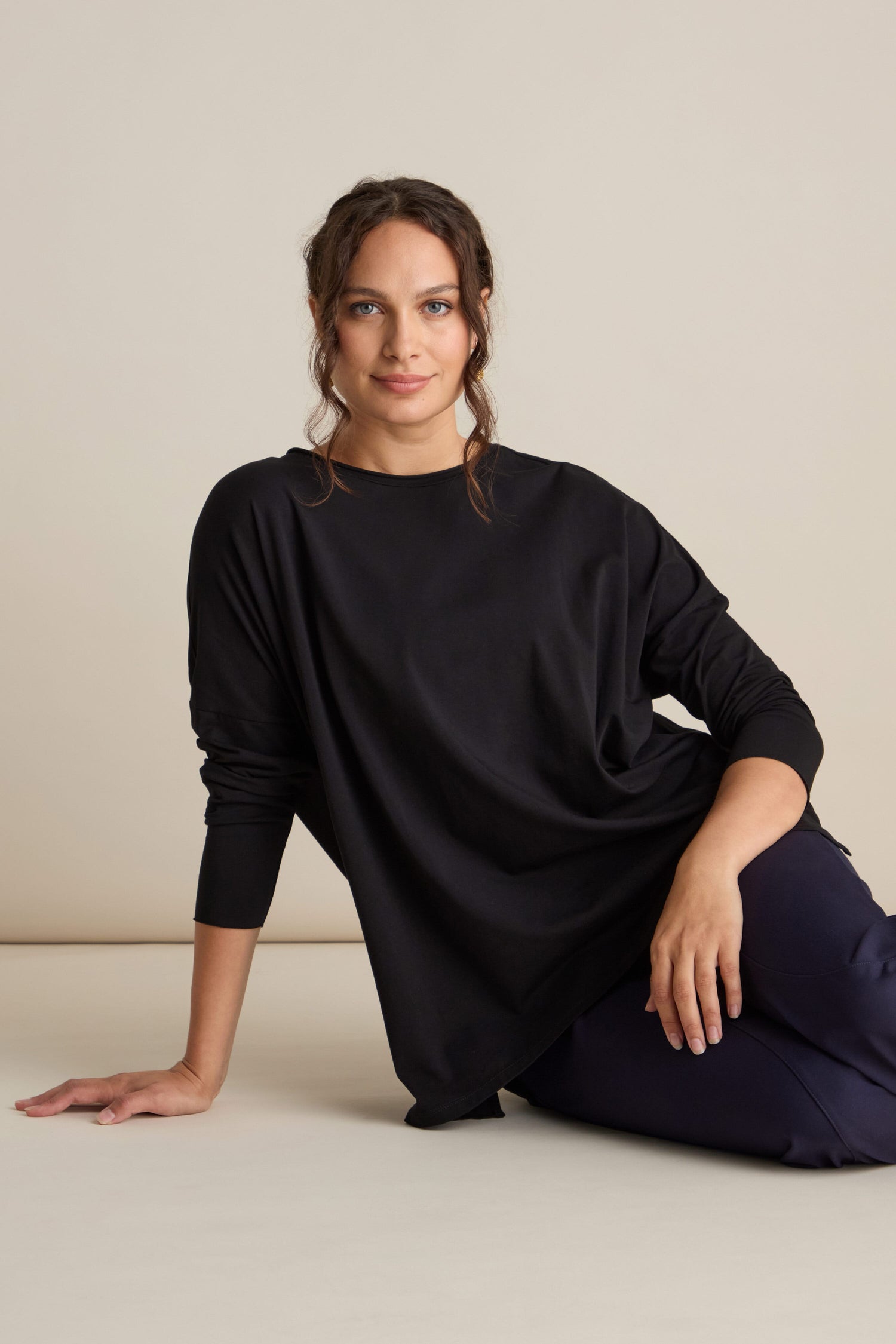 A person with long hair is seated on the floor against a plain background, smiling slightly while leaning on one hand. They are wearing dark pants and the Cotton Jersey Relaxed Top in black, adding to their effortless style.