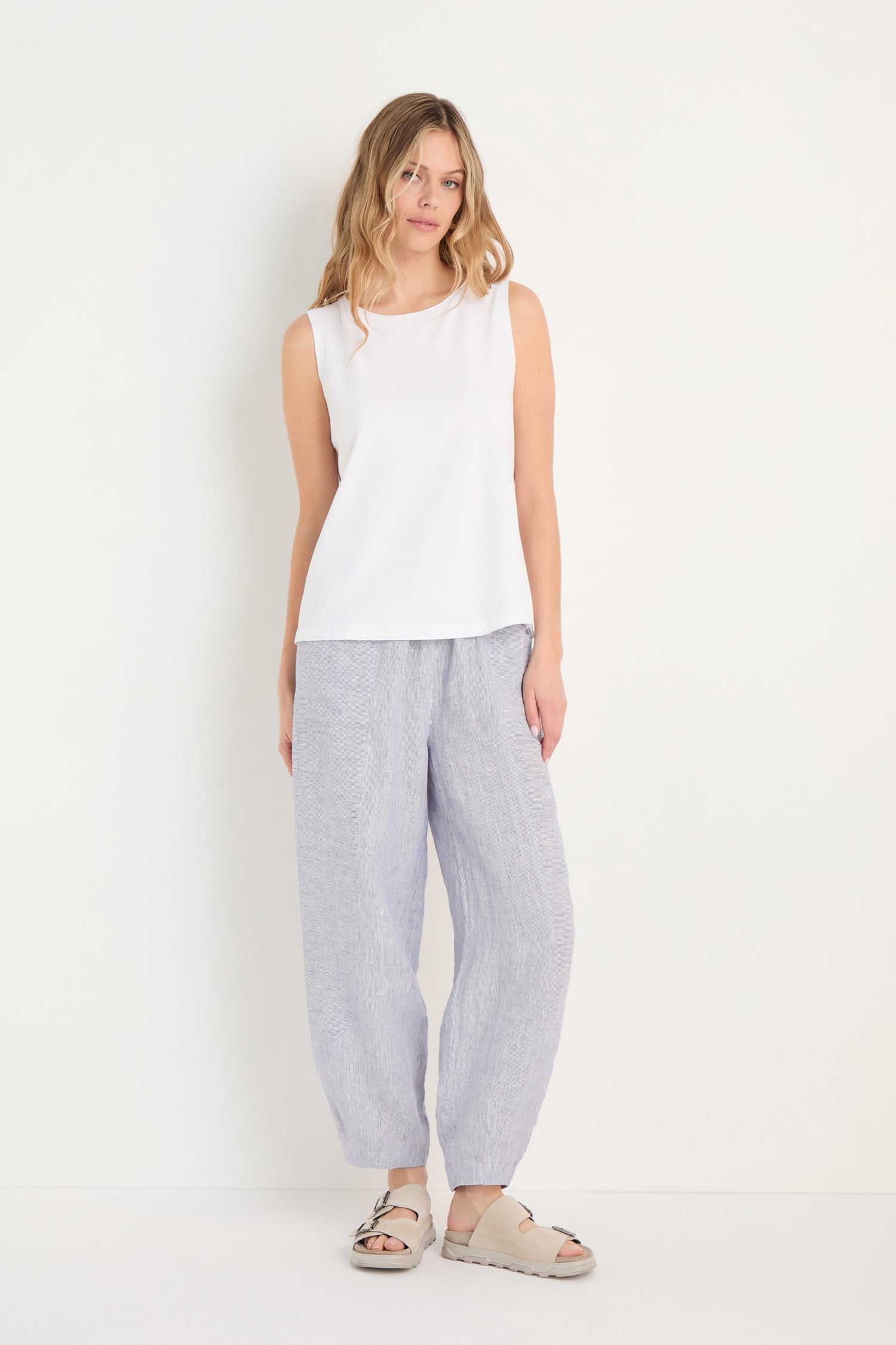 Woman standing against a white background, wearing the Cotton Jersey Boxy Vest in white, light gray loose linen pants, and beige sandals.