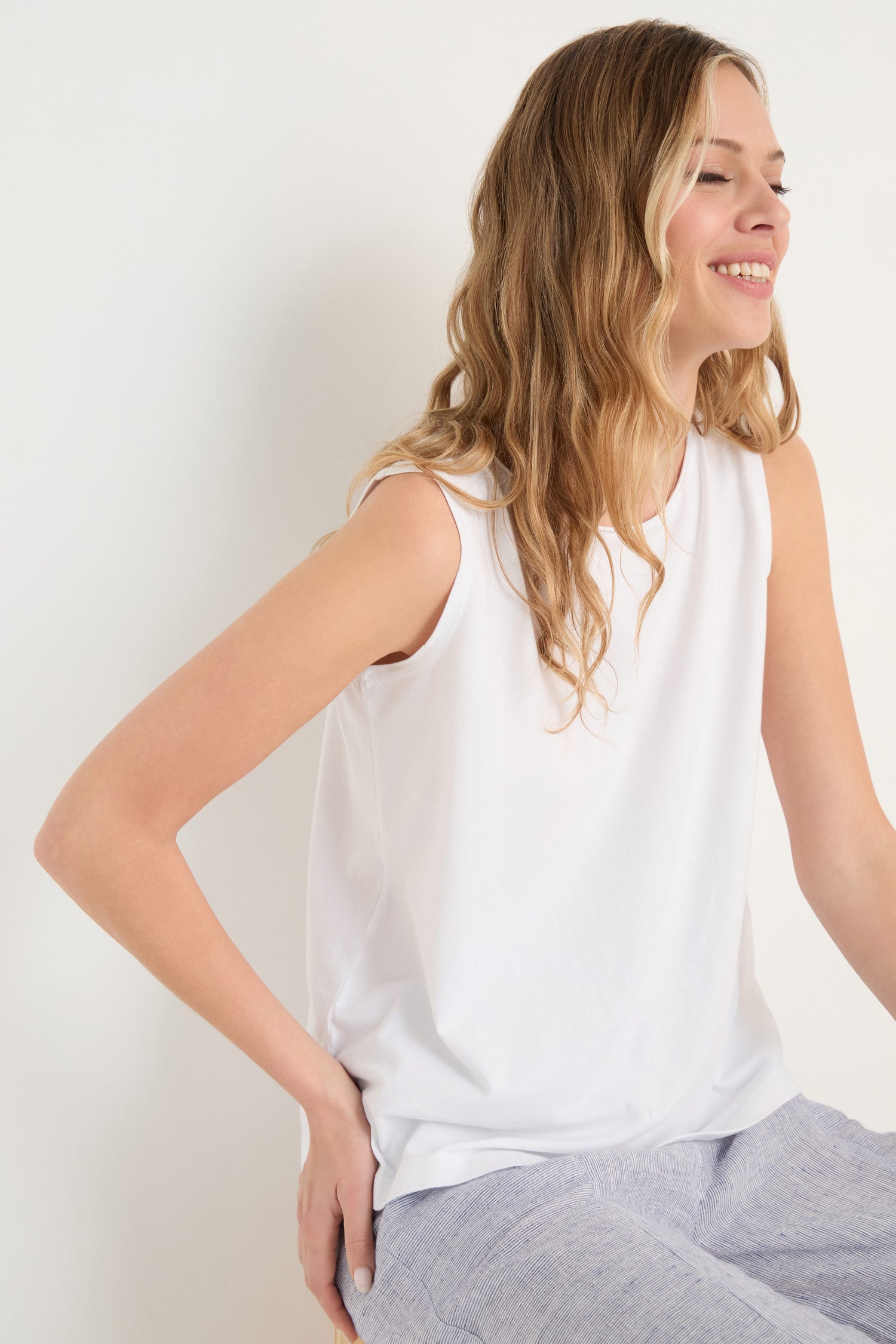 A woman with long blonde hair smiles, seated against a plain background, wearing the Cotton Jersey Boxy Vest and light gray pants.