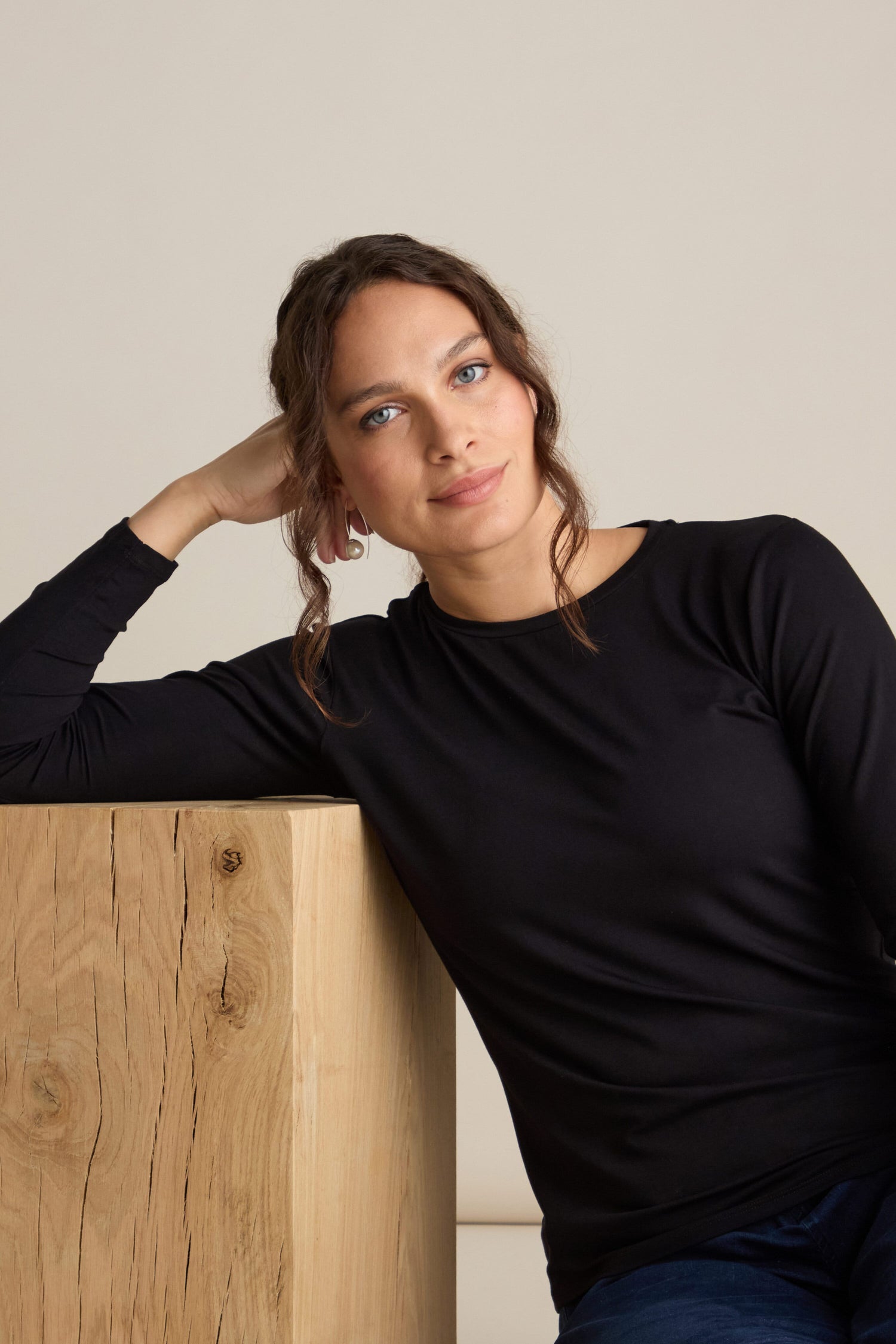 A person with long hair leans on a wooden surface, wearing a Viscose Jersey Relaxed Top with long sleeves in black, a true wardrobe staple, and looks directly at the camera.