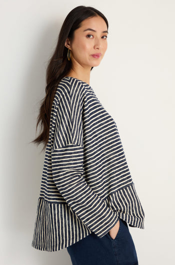 A woman stands sideways against a white wall, wearing the Textured Stripe Jersey Boxy Top in navy and white with dark jeans and her hands in her pockets.