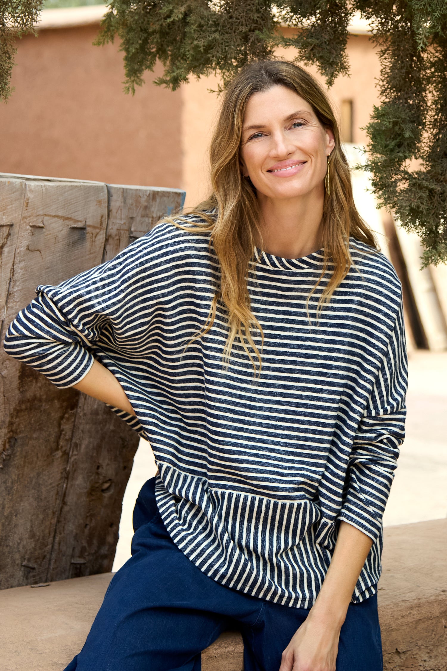 A woman with long, wavy hair sits outdoors, smiling at the camera in a Textured Stripe Jersey Boxy Top and navy pants.