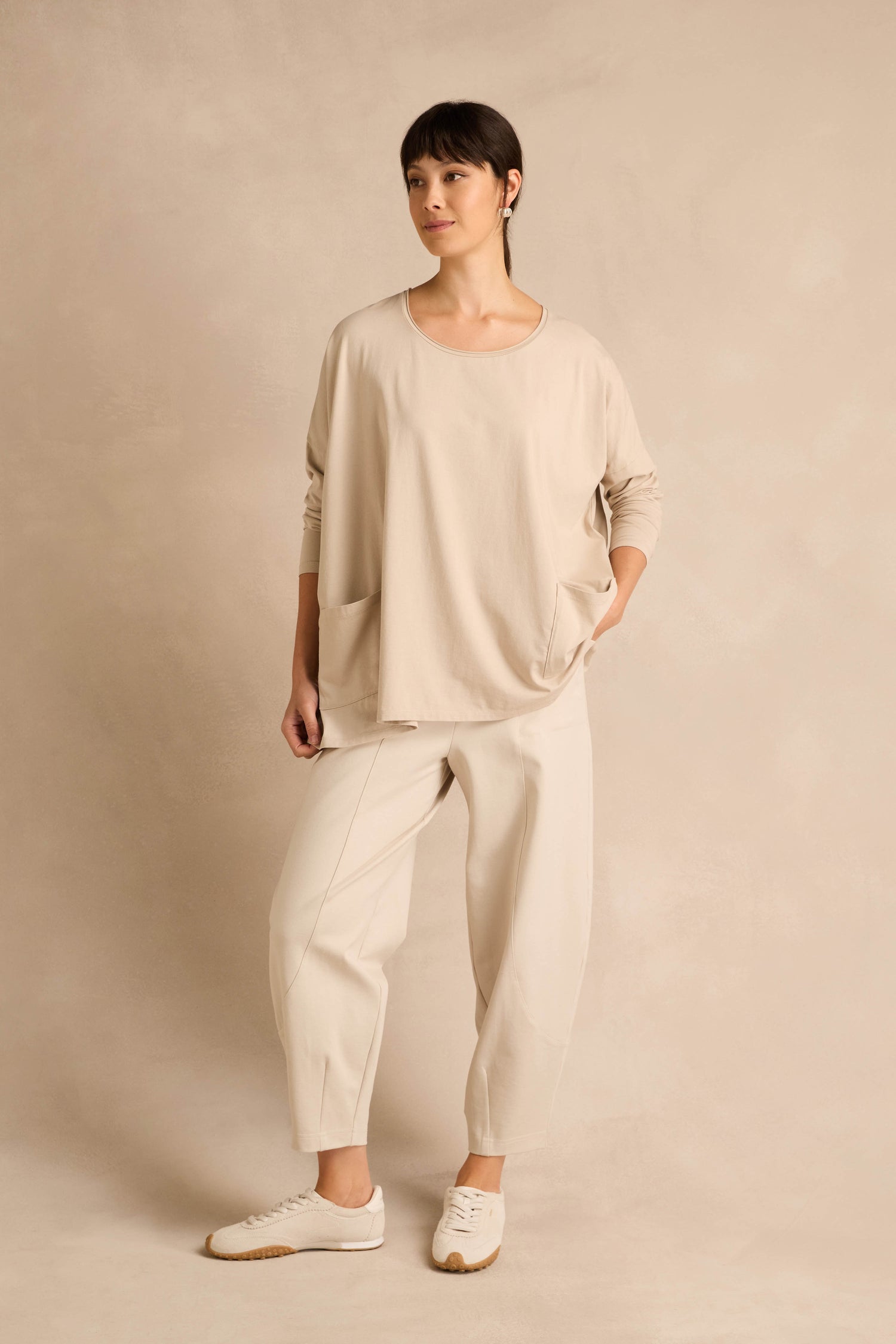 A woman stands against a neutral background wearing the Ecru Cotton Jersey Pocket Top, paired with beige pants and beige sneakers.