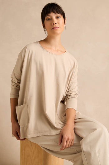 Wearing the Ecru Cotton Jersey Pocket Top and matching pants, a woman sits on a wooden stool against a neutral background, looking at the camera with a relaxed expression.