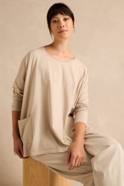 Wearing the Ecru Cotton Jersey Pocket Top and matching pants, a woman sits on a wooden stool against a neutral background, looking at the camera with a relaxed expression.