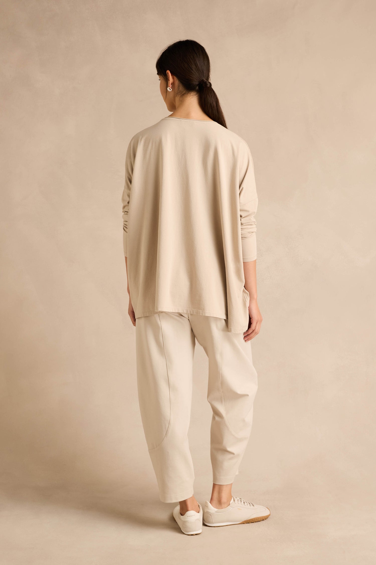 A person stands facing away, wearing the loose Ecru Cotton Jersey Pocket Top, matching pants, and white sneakers, with their hair tied back in a ponytail against a plain beige background.