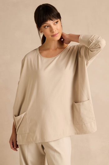 A woman wears the Ecru Cotton Jersey Pocket Top—an oversized, relaxed shirt in beige with two front pockets—paired with matching beige pants, standing against a beige wall with her hand resting on her neck.