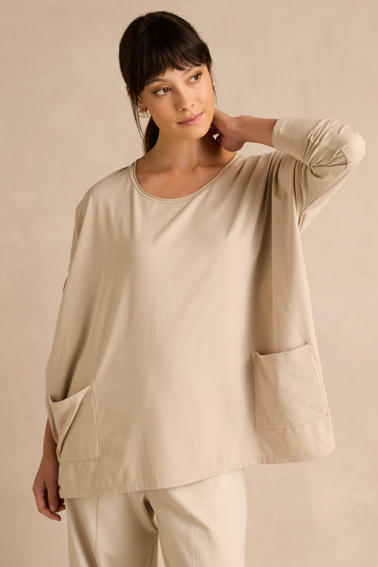 A woman wears the Ecru Cotton Jersey Pocket Top—an oversized, relaxed shirt in beige with two front pockets—paired with matching beige pants, standing against a beige wall with her hand resting on her neck.