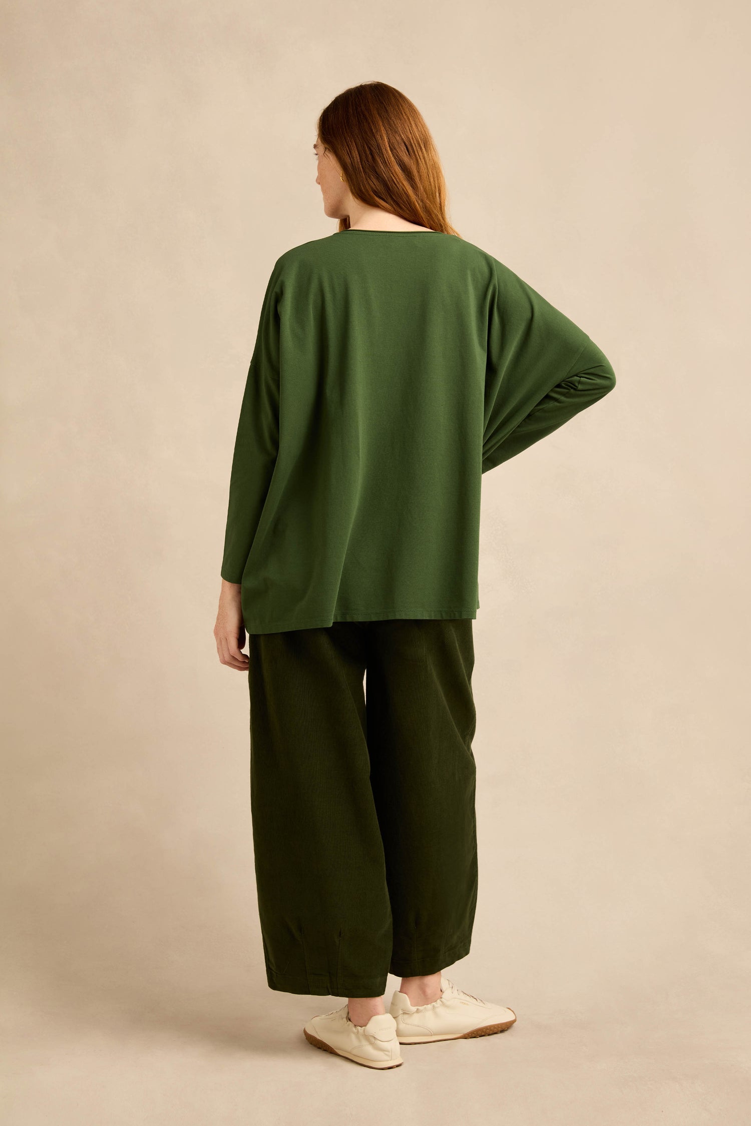 A person is seen from behind, wearing the Forest Cotton Jersey Pocket Top in a loose, boxy silhouette with wide-leg dark green pants and white shoes, set against a neutral beige backdrop.