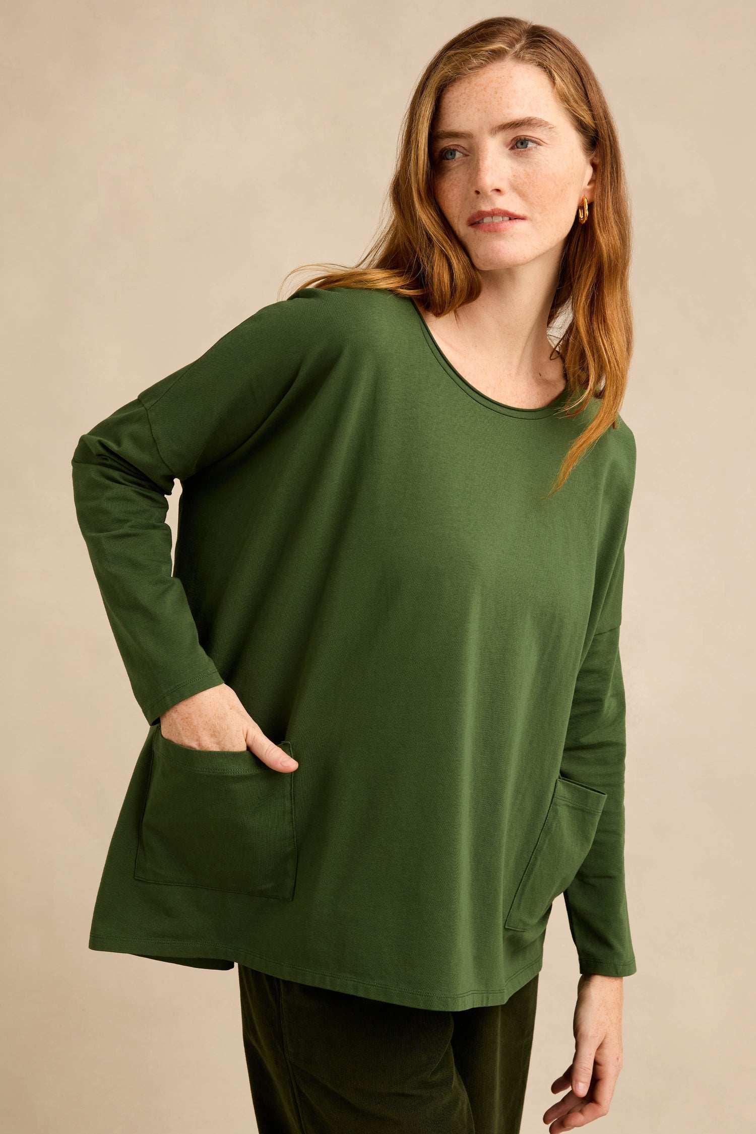 A woman stands against a beige background wearing the Forest Cotton Jersey Pocket Top, a relaxed, oversized, long-sleeved shirt in dark green with two front pockets and matching pants.