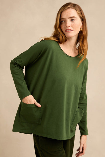 A woman stands against a beige background wearing the Forest Cotton Jersey Pocket Top, a relaxed, oversized, long-sleeved shirt in dark green with two front pockets and matching pants.