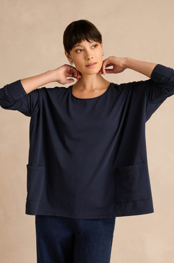 A woman stands against a beige background, wearing the Navy Cotton Jersey Pocket Top with front pockets and dark pants, her hands held near her neck.