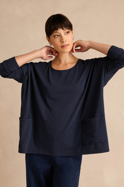 A woman stands against a beige background, wearing the Navy Cotton Jersey Pocket Top with front pockets and dark pants, her hands held near her neck.