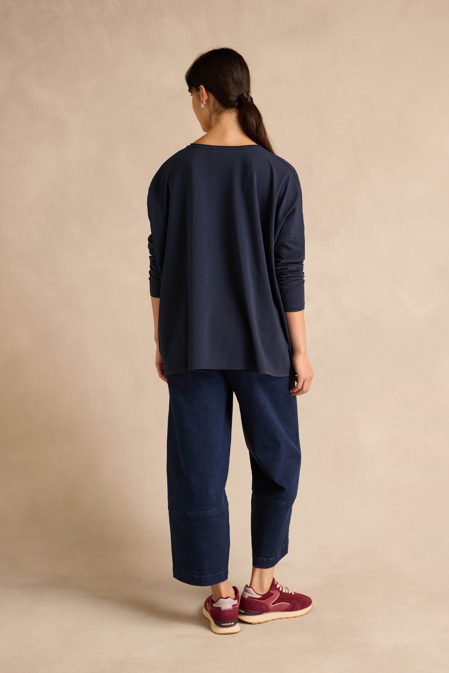 A person with long brown hair in a ponytail wears the Navy Cotton Jersey Pocket Top styled oversized, along with wide-leg denim pants and burgundy sneakers, standing with their back to the camera against a beige background.