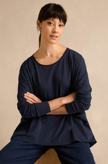 A woman with straight dark hair, wearing the Navy Cotton Jersey Pocket Top and jeans, sits with her arms crossed against a plain beige background.