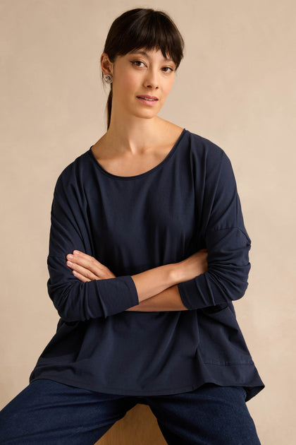 A woman with straight dark hair, wearing the Navy Cotton Jersey Pocket Top and jeans, sits with her arms crossed against a plain beige background.