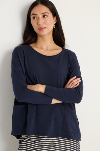 A woman with long dark hair stands against a light wall, arms crossed, wearing the Cotton Jersey Boxy Pocket Top in navy blue with an oversized fit and a striped skirt.