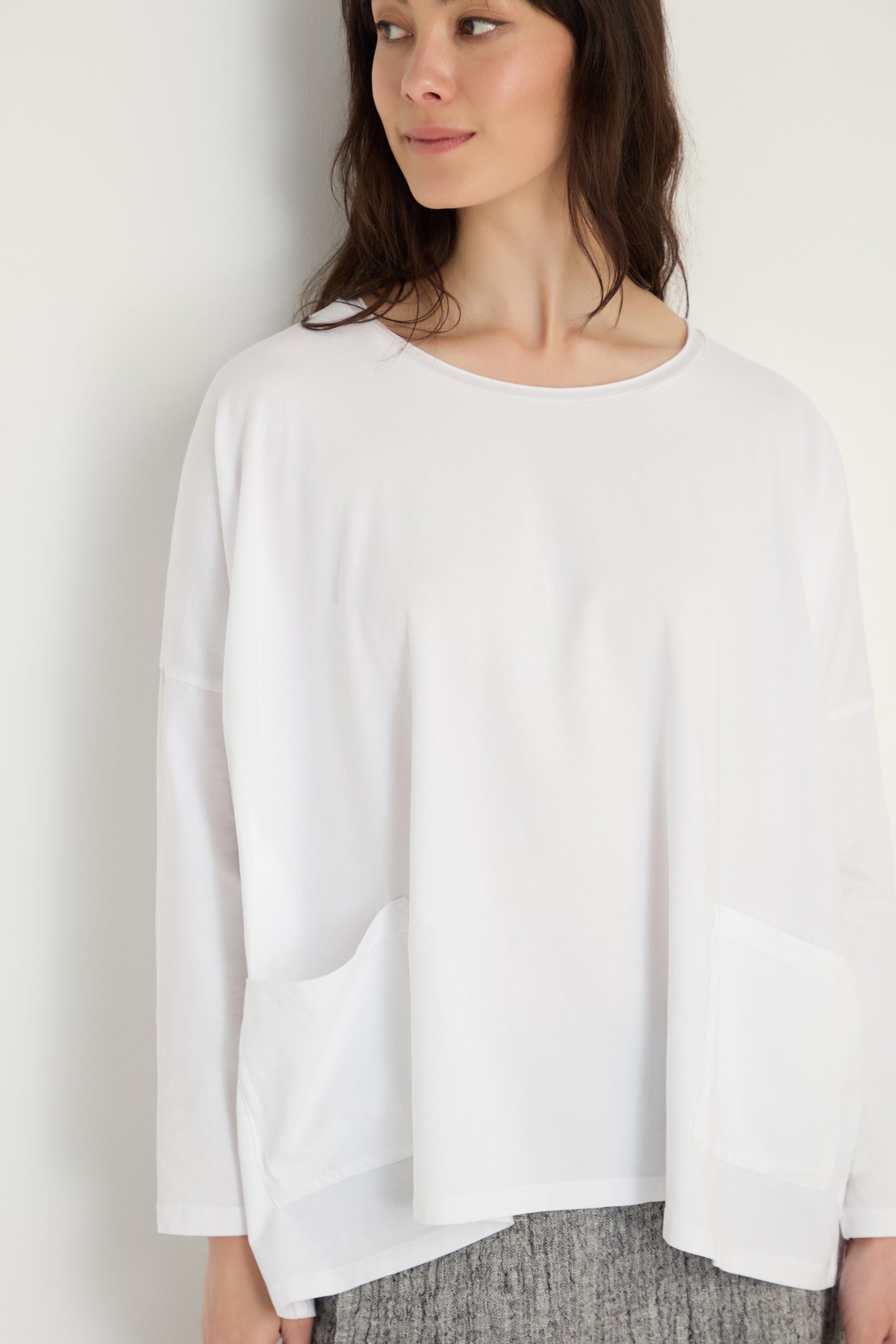 A woman wears the Cotton Jersey Boxy Pocket Top—a loose-fitting white long-sleeve with two front pockets—paired with gray textured pants, standing against a plain white background.